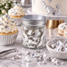 A clear glass jar filled with silver leaf confetti sits on a marble surface, styled with a decorated cupcake, macarons, and a cake in the background.