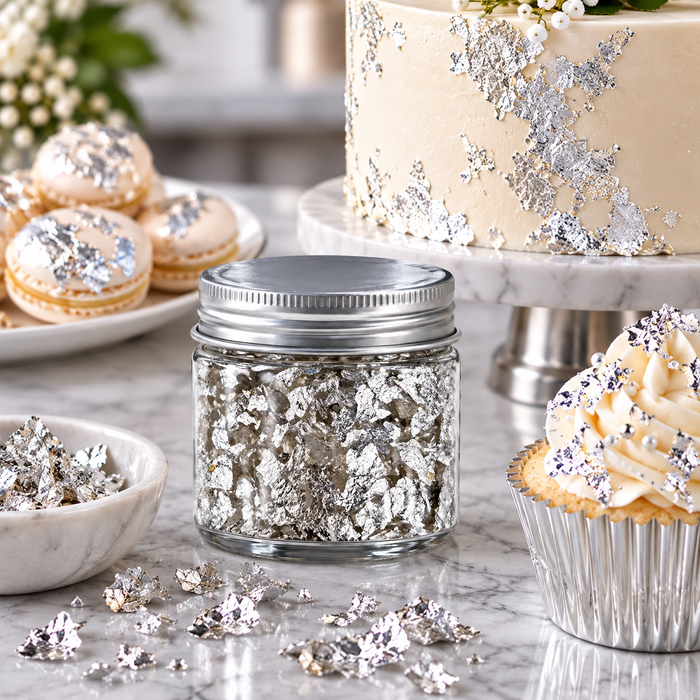 A small clear glass jar filled with silver leaf flakes and topped with a silver lid, styled on a marble surface with cupcakes, macarons, and a decorated cake in the background.