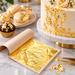 Gold leaf flake sheets displayed on parchment paper with a decorated cream cake, macarons, and a chocolate cupcake in the background on a marble surface.