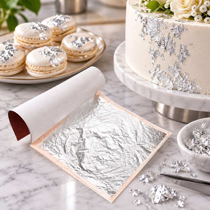 Silver leaf sheeting paper shown unrolled on a marble surface, with scattered crumpled silver leaf pieces nearby, a plate of macarons, and a decorated cake in the background.